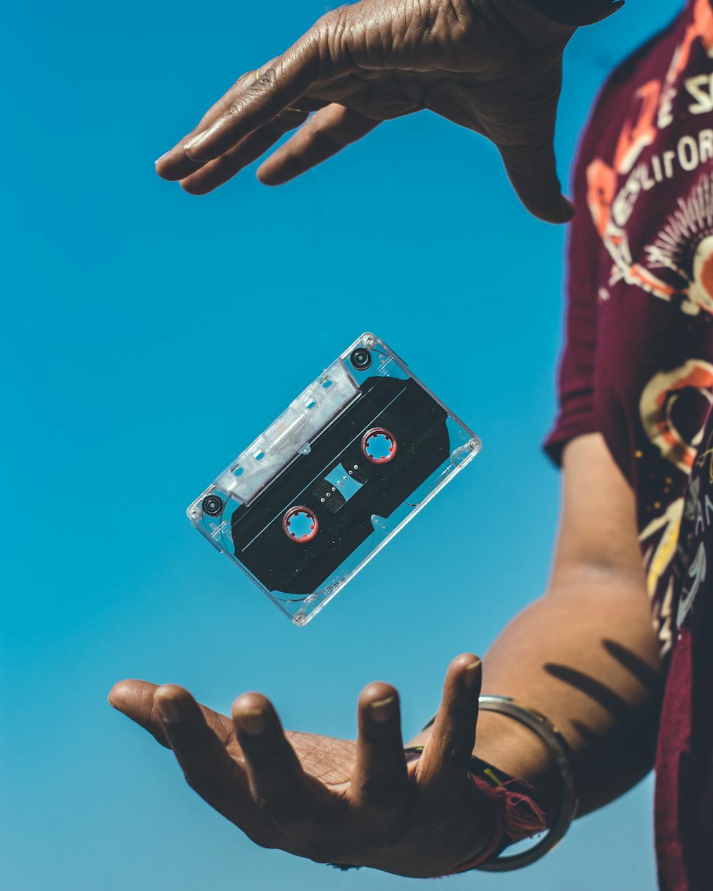 A retro cassette tape floating between hands against a clear blue sky, evoking nostalgia.